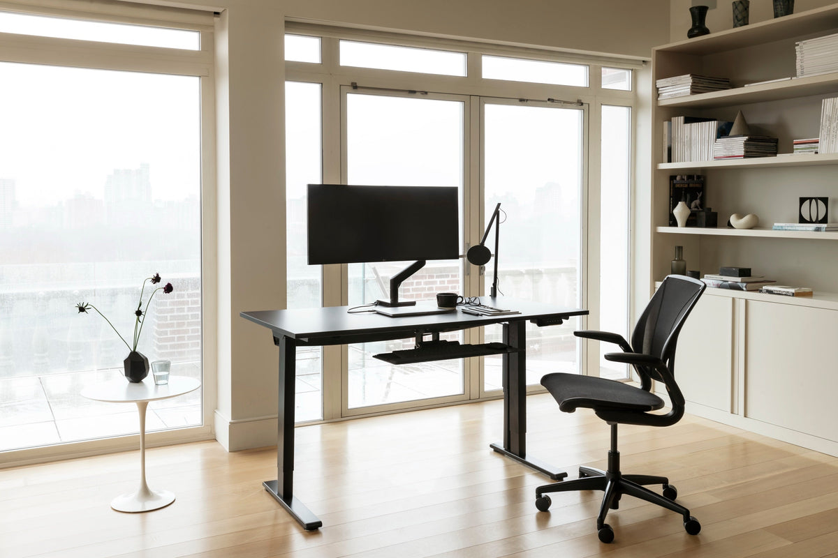 A modern all-black Humanscale office setup featuring a Liberty Ocean chair in black pinstripe mesh, a black eFloat Go 2.0 desk, a black M2 Pro monitor arm, and a black Nova LED lamp near a window