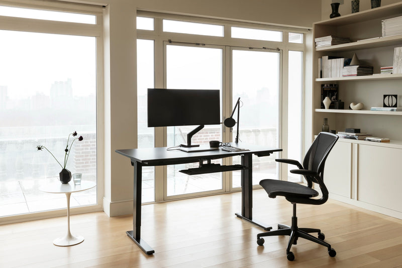 A modern all-black Humanscale office setup featuring a Liberty Ocean chair in black pinstripe mesh, a black eFloat Go 2.0 desk, a black M2 Pro monitor arm, and a black Nova LED lamp near a window