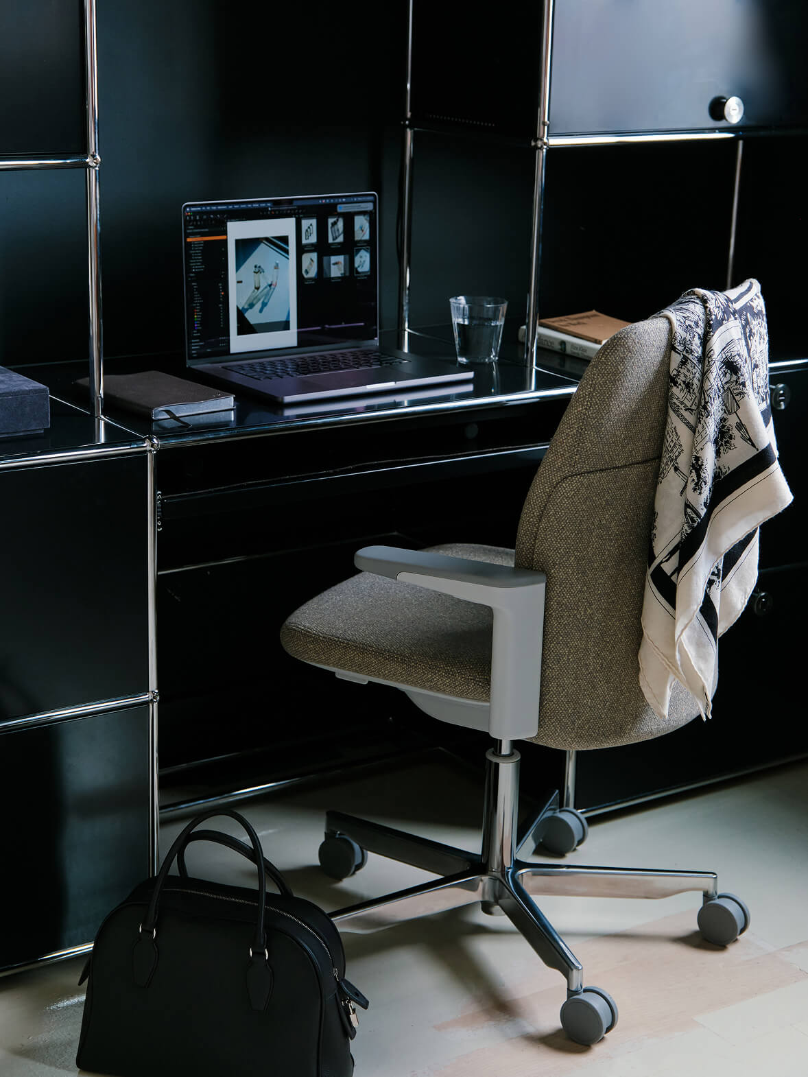 Modern office desk with laptop, chair, and personal items in a dark moody room with bookshelf. 