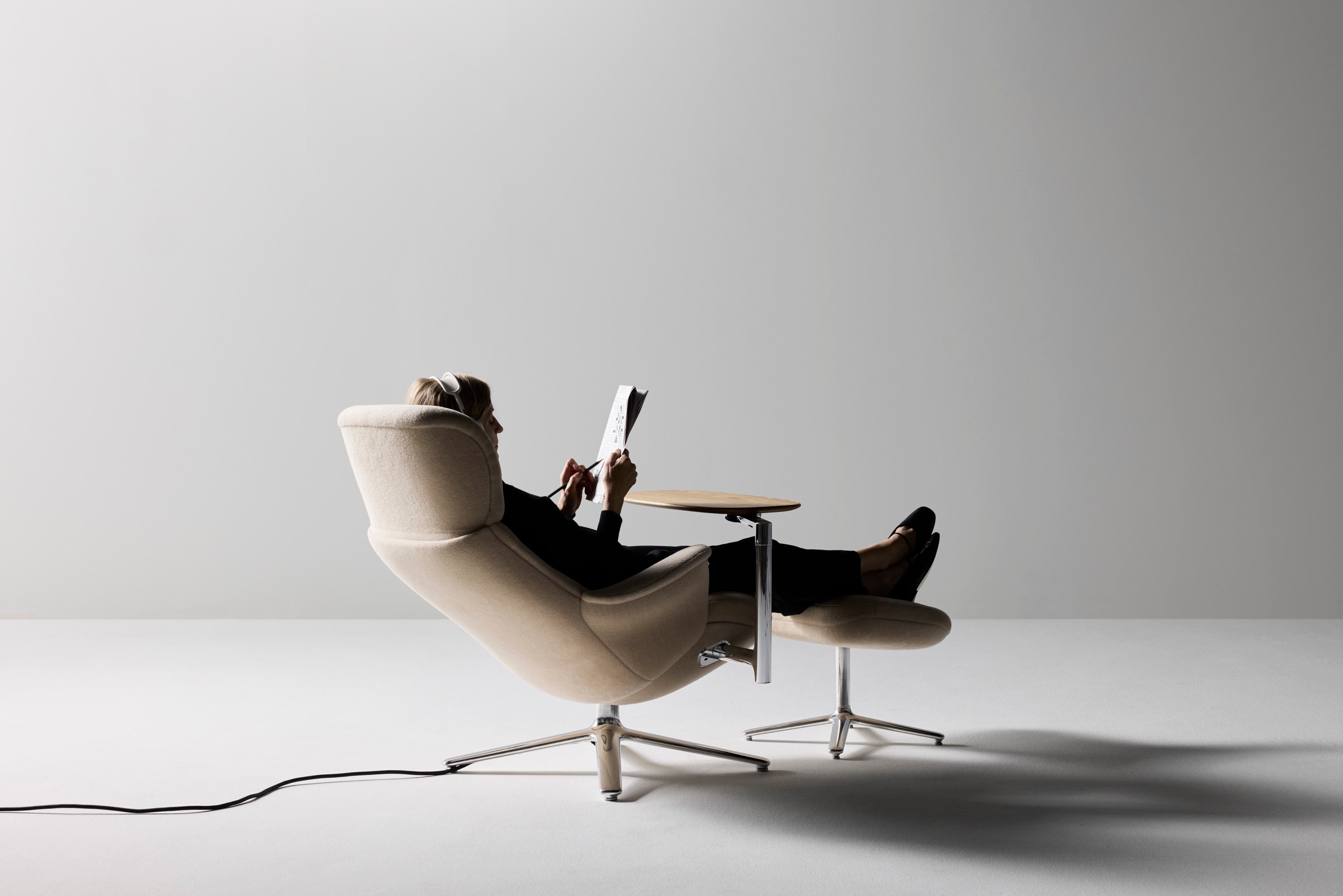 A rear diagonal view of a person relaxing and reading a book in a beige Humanscale Diffrient Lounge chair with a matching ottoman and small side table in a cozy home lounge setting