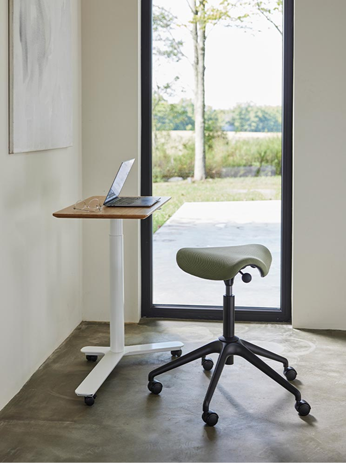 A Humanscale Freedom Active Pony Stool with a black frame and Driftwood cushion seat next to a Float Mini adjustable table with a white base and wood top surface