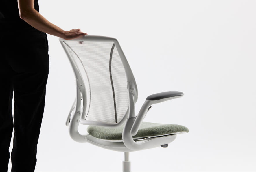 A person stands behind a white Humanscale Liberty task chair, pushing it with their hand resting on the Pinstripe Silver mesh backrest, showcasing the chair's green textile seat and all-white frame, base, and trim