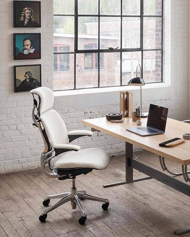 A bright loft-style office setup featuring a white Humanscale Freedom Ocean task chair and an eFloat Go standing desk with a laptop and lamp against a white brick wall background