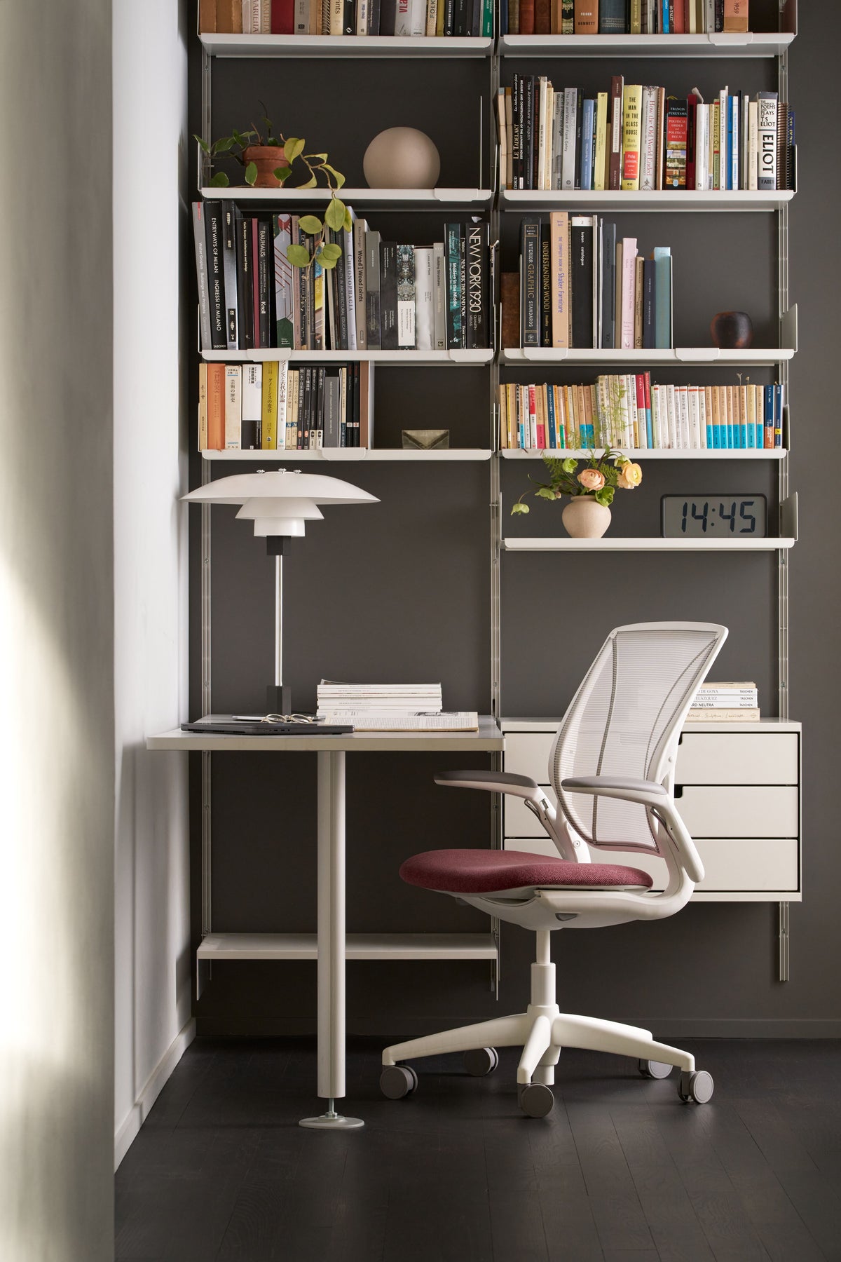 Portrait view of a stylish home office setup with a Humanscale World Upholstered chair, white mesh back, red seat cushion, and white frame, in a dark grey room