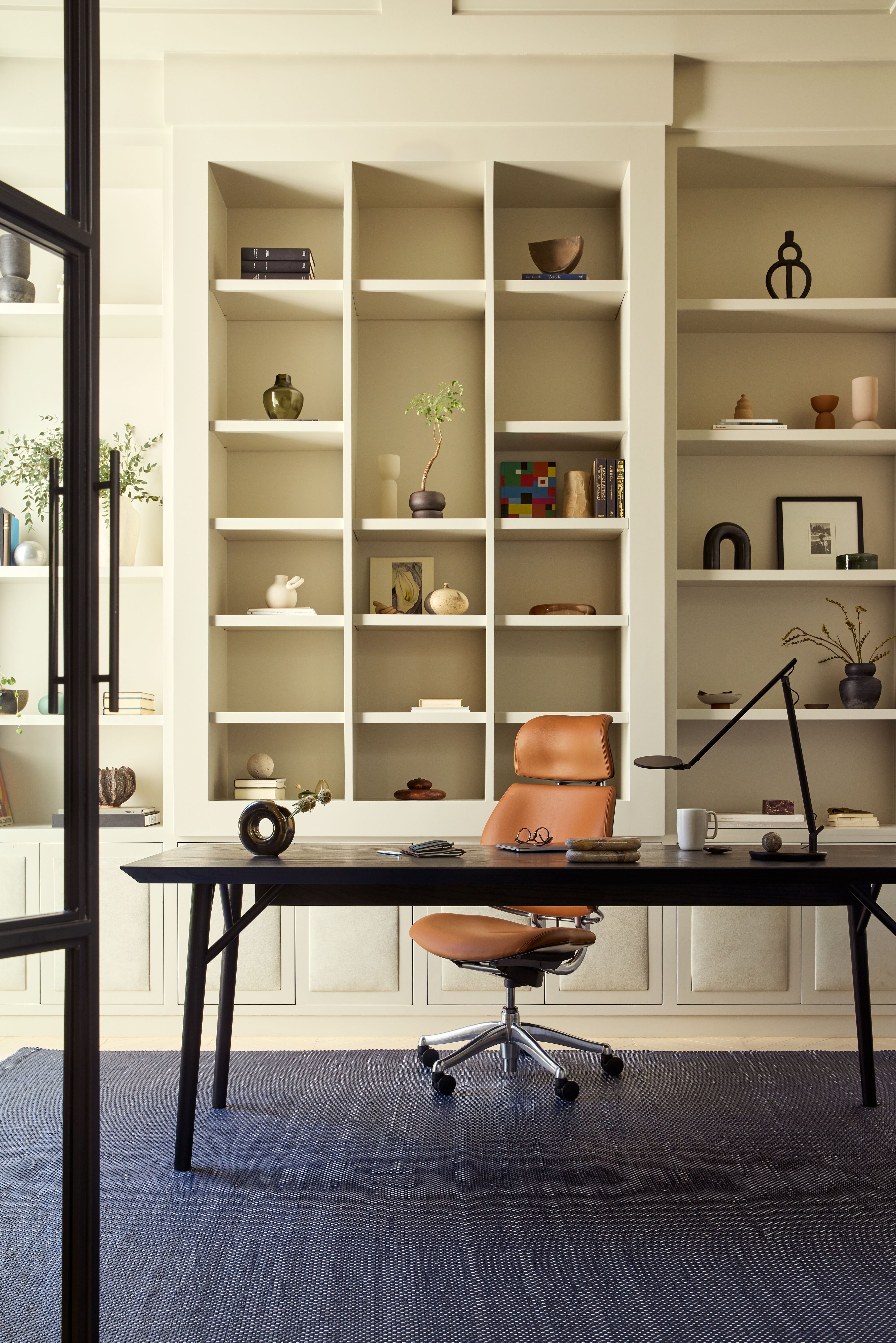 A portrait front view of a luxury home office featuring a tan Saddle Leather Humanscale Freedom Ocean chair with headrest and a black Nova LED desk lamp on a wooden desk with a bookshelf in the background