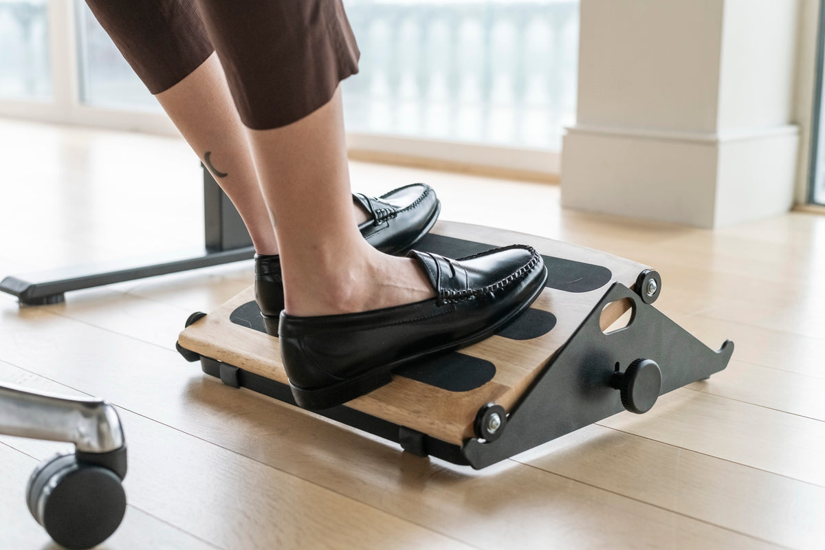 Top-down lifestyle view of a person using a black Humanscale FR300 ergonomic foot rocker with a solid hardwood platform and non-skid pads to encourage leg movement