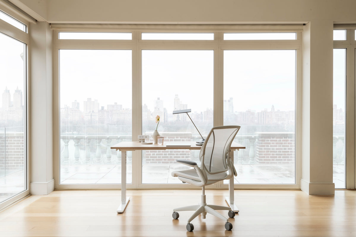 White and silver Humanscale Diffrient World chair with a solid oak eFloat Go 2.0 desk and a silver Horizon 2.0 lamp in a modern office with city views