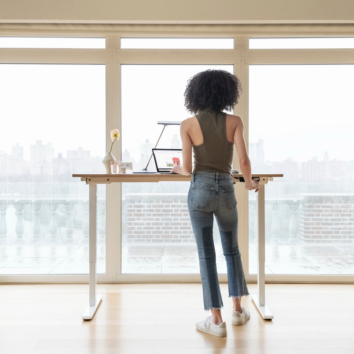 Woman standing at a Humanscale eFloat Go 2.0 solid oak desk with a silver Horizon 2.0 task lamp in front of a city skyline view