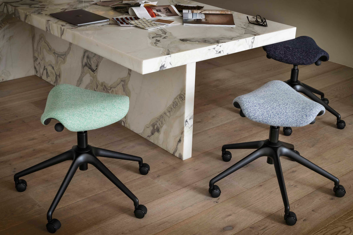 Three Humanscale Freedom Active Pony stools in black, light green, and light blue upholstery arranged around a marble-topped table, highlighting the seven-degree movement for collaborative environments