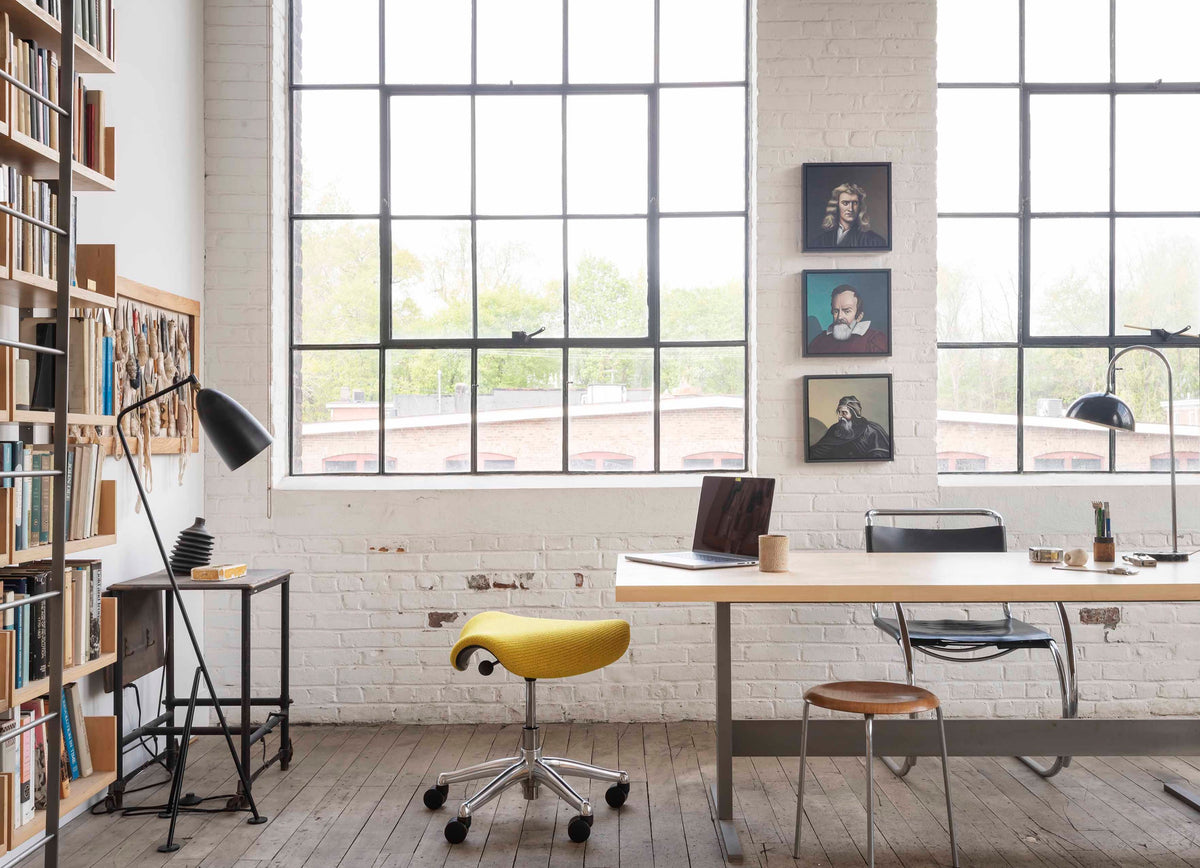A horizontal view of a mustard yellow Humanscale Freedom Saddle stool with a polished aluminum frame and graphite trim in a sunlit modern office featuring large windows, a desk with a laptop, and a pedestal lamp