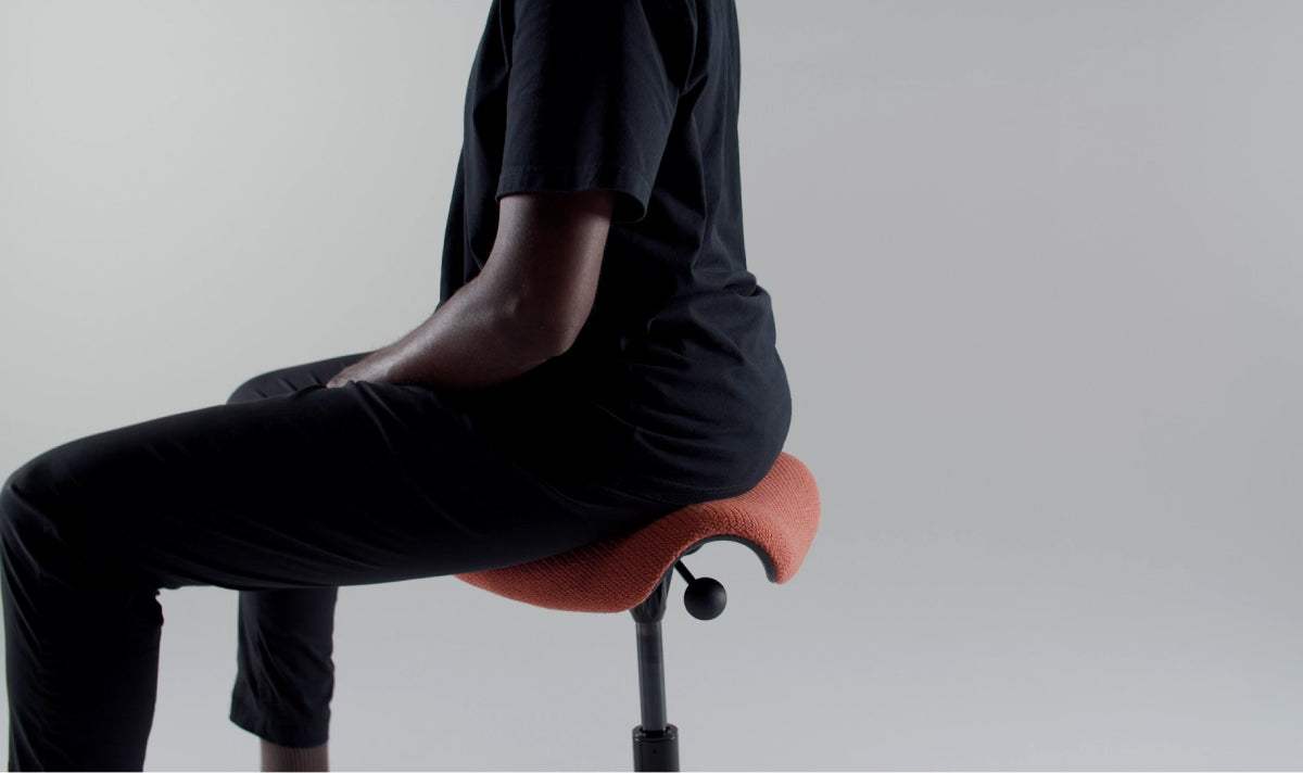 A person in black clothing demonstrating active sitting on an orange Humanscale Saddle Stool, showing how the triangular cushion opens the hips and encourages a healthy S-curve spine posture