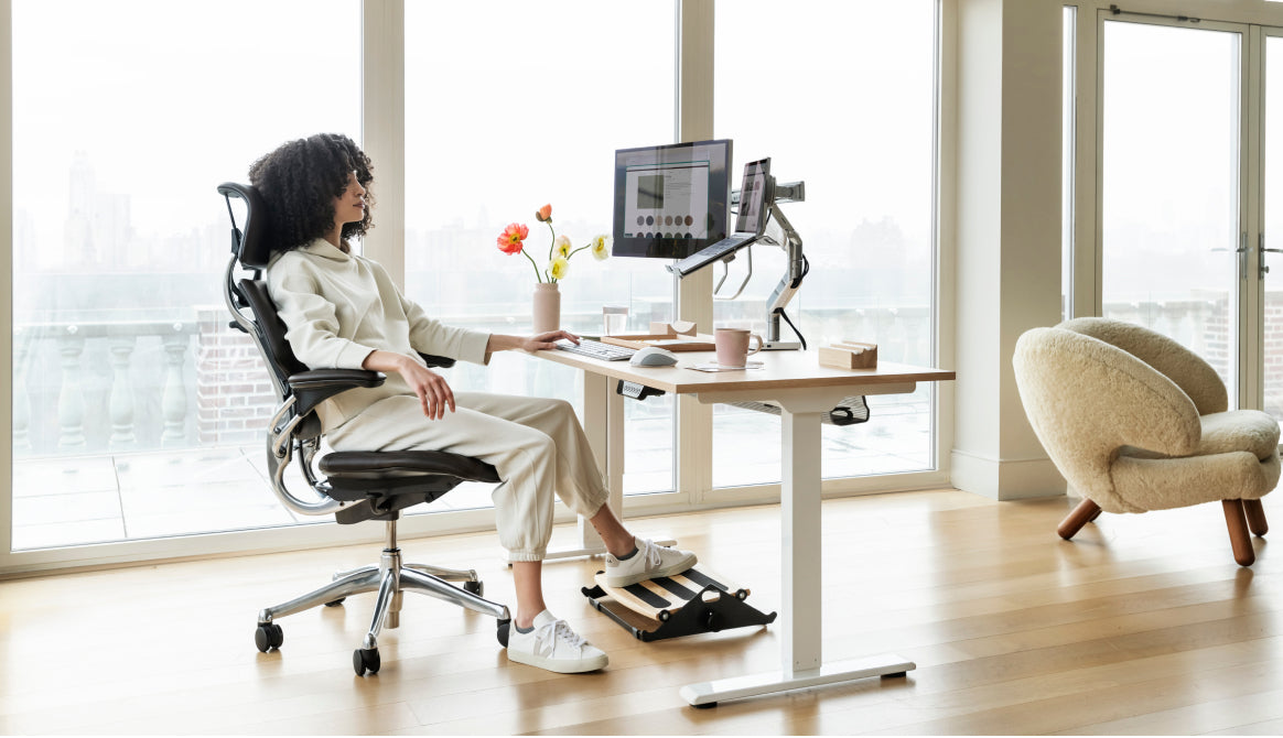 A person working at a Humanscale eFloat Go 2.0 height-adjustable desk on a Freedom 25 Signature Edition chair, featuring a monitor arm with notebook holder, universal accessory bracket, and an FR300 foot rocker in a bright office with large windows