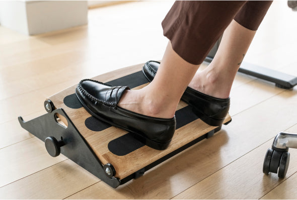 A person using the Humanscale FR300 Foot Rocker with a solid wood platform and black adjustable frame on a light wood floor to improve office ergonomics