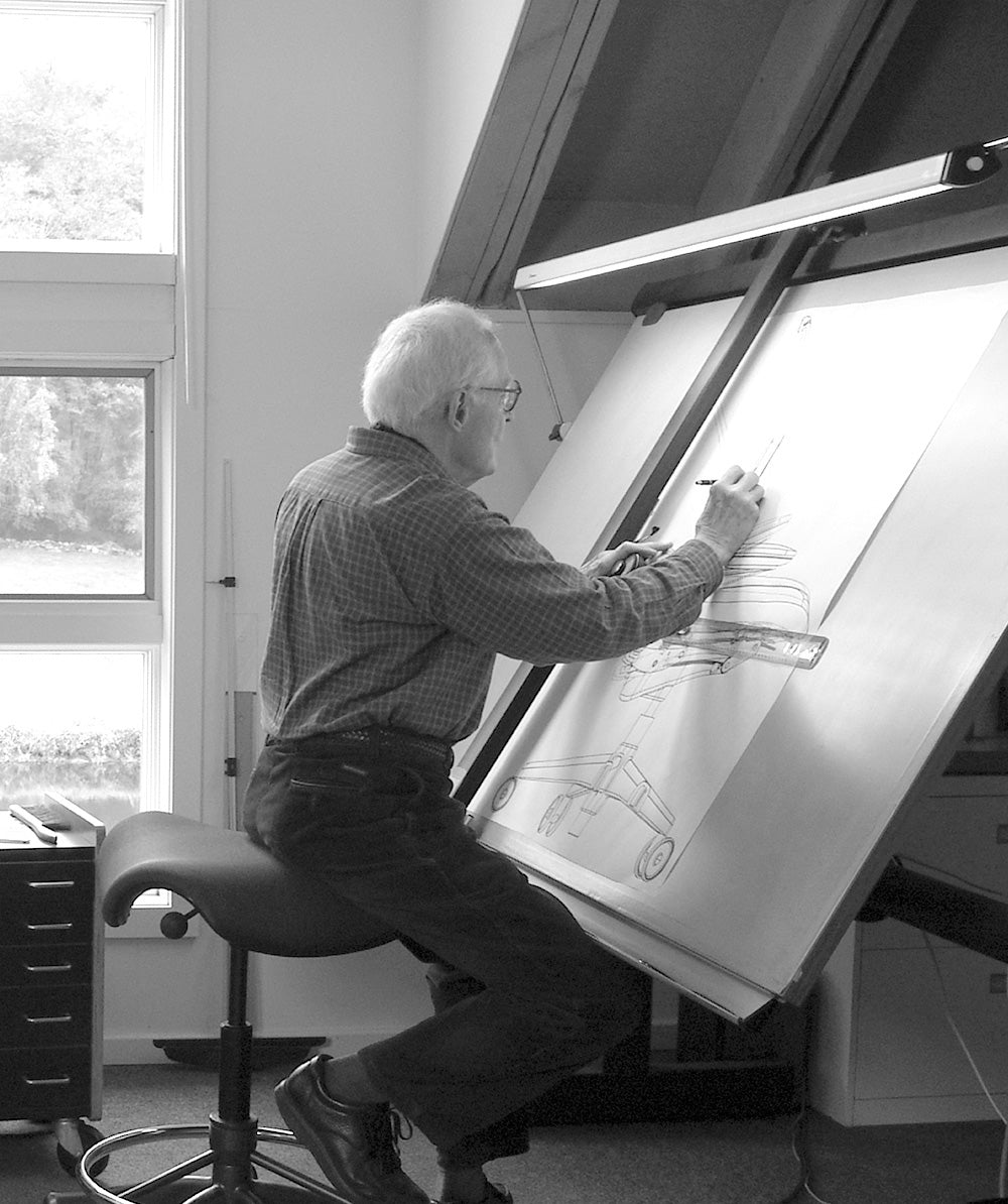 Industrial designer Niels Diffrient seated on a Humanscale Freedom Saddle stool with a high cylinder and footring, working at a large drafting table on a chair design sketch
