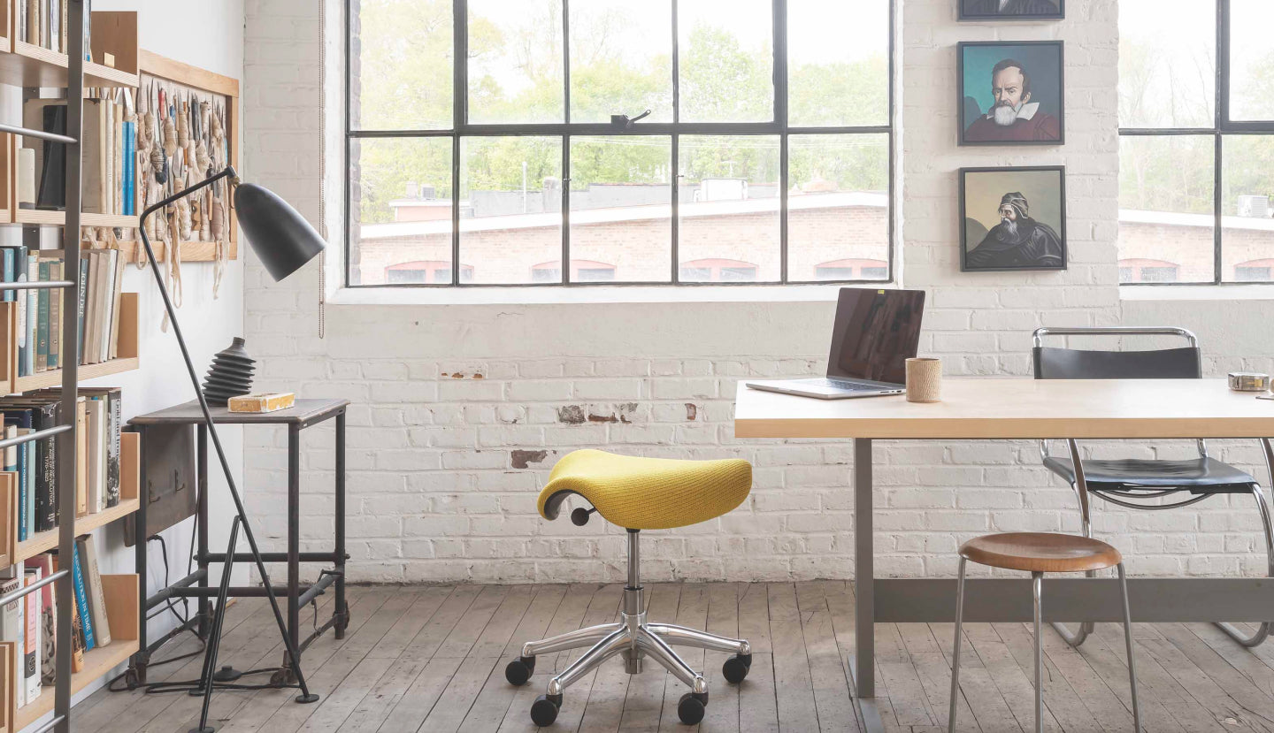 A horizontal view of a mustard yellow Humanscale Freedom Saddle stool with a polished aluminum frame and graphite trim in a sunlit modern office featuring large windows, a desk with a laptop, and a pedestal lamp