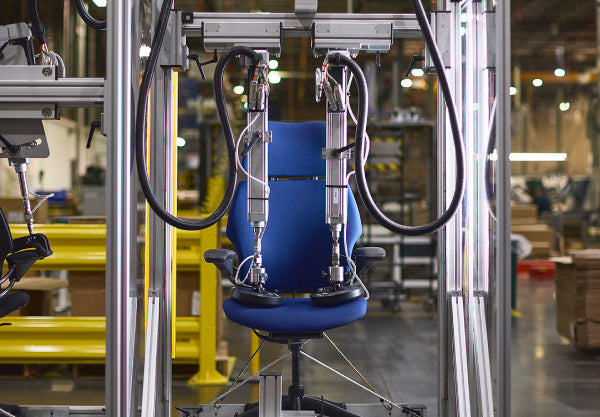 An automated machine performing durability and pressure testing on a blue Humanscale Freedom Ocean sustainable task chair with a black frame and trim in a factory setting