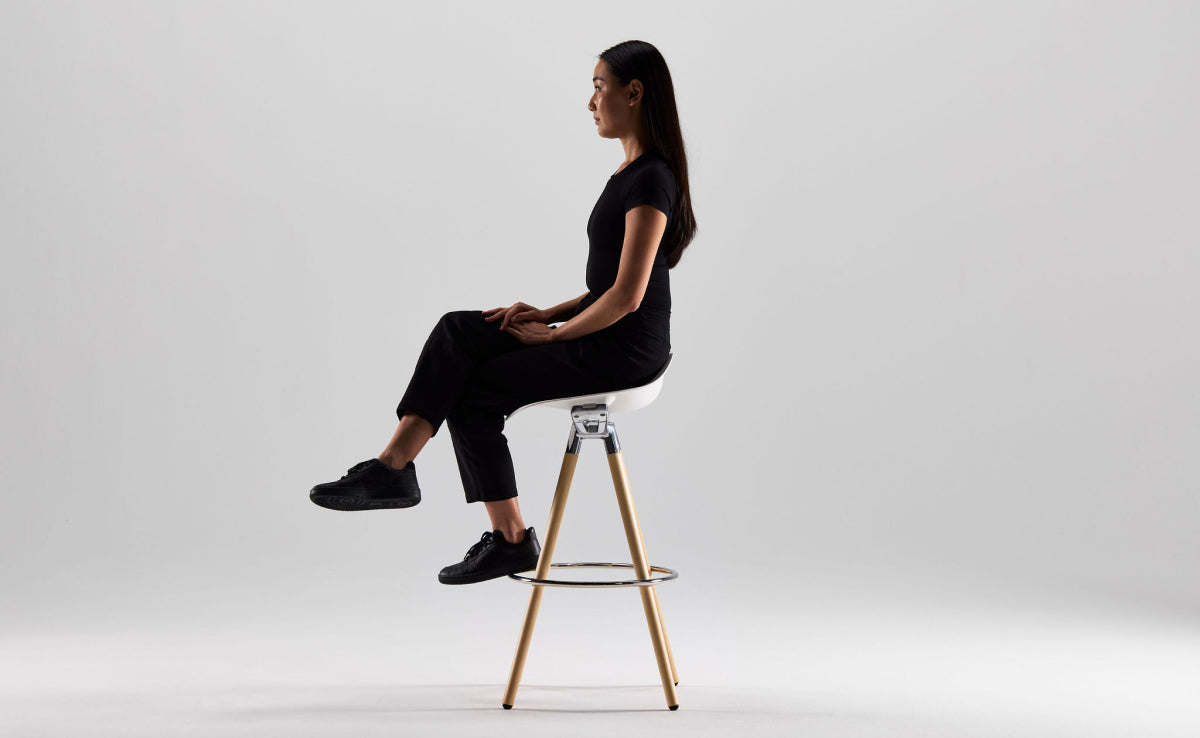 A side view of a woman sitting upright with crossed legs on a white Humanscale Sedeo stool with bar-height polished aluminum legs and a metal footrest ring against a plain light gray background