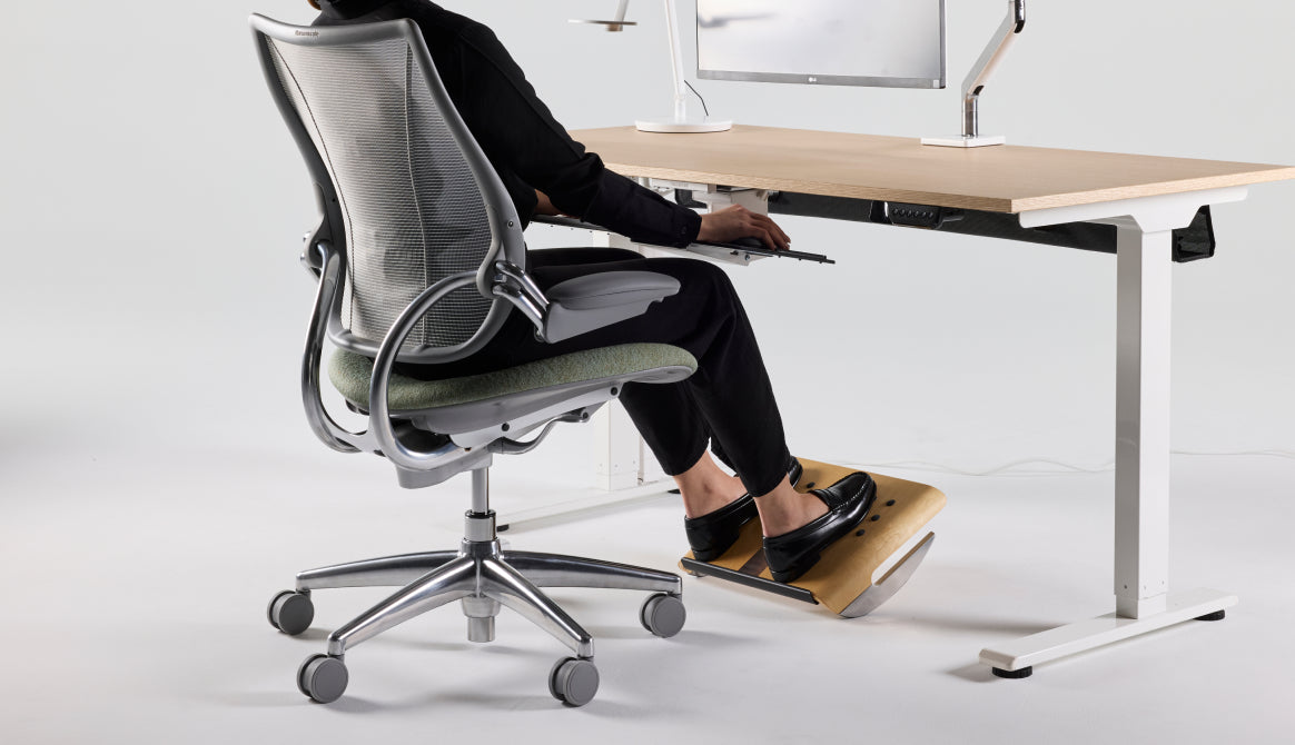 Lifestyle view of a person working at an eFloat Go 2.0 standing desk, seated in a Liberty Task chair with silver mesh, and using a FR500 ergonomic foot rocker for active posture
