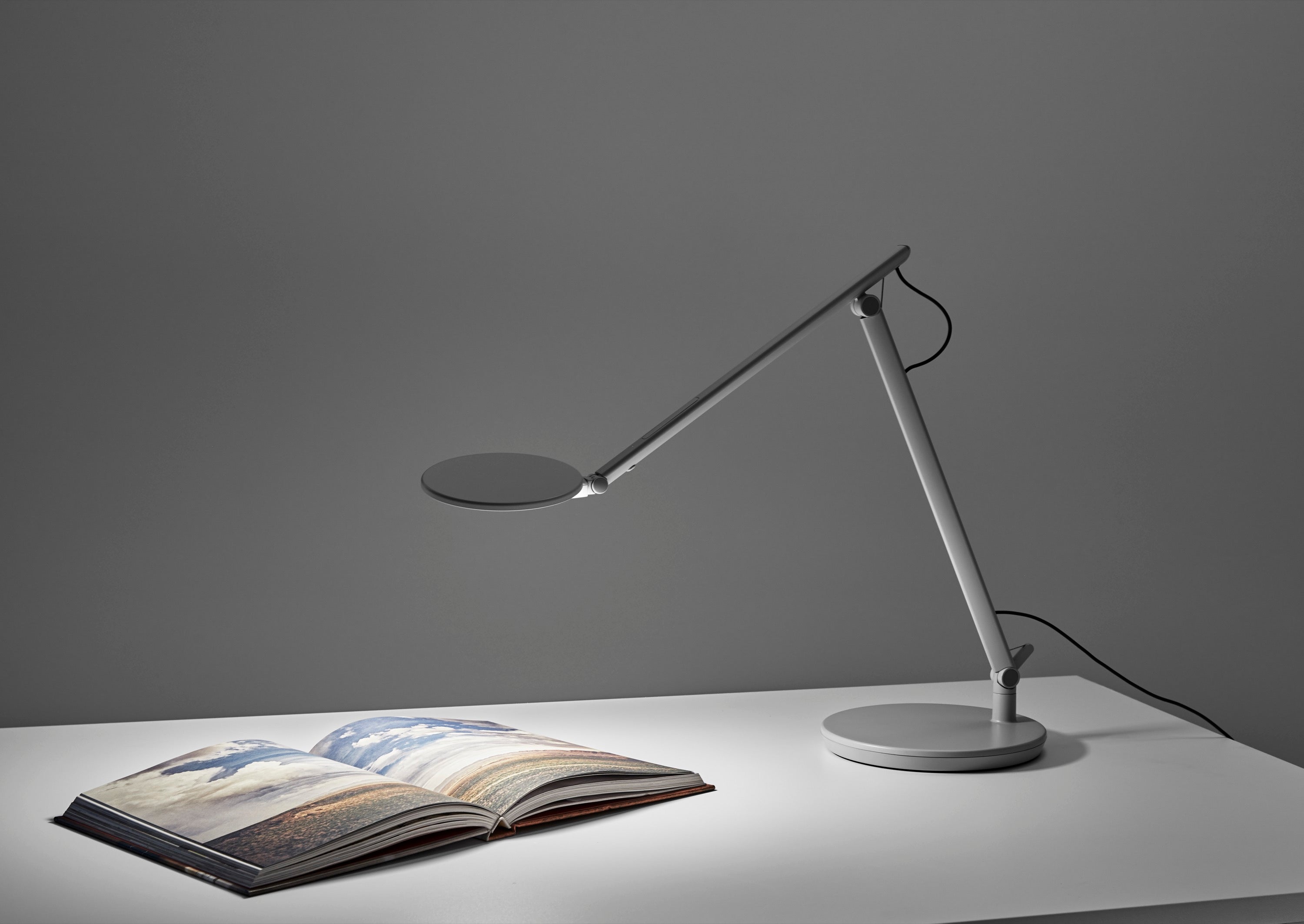 A silver Humanscale Nova LED task light with a desktop base illuminating an open photography book with cool white light on a white desk against a gray wall