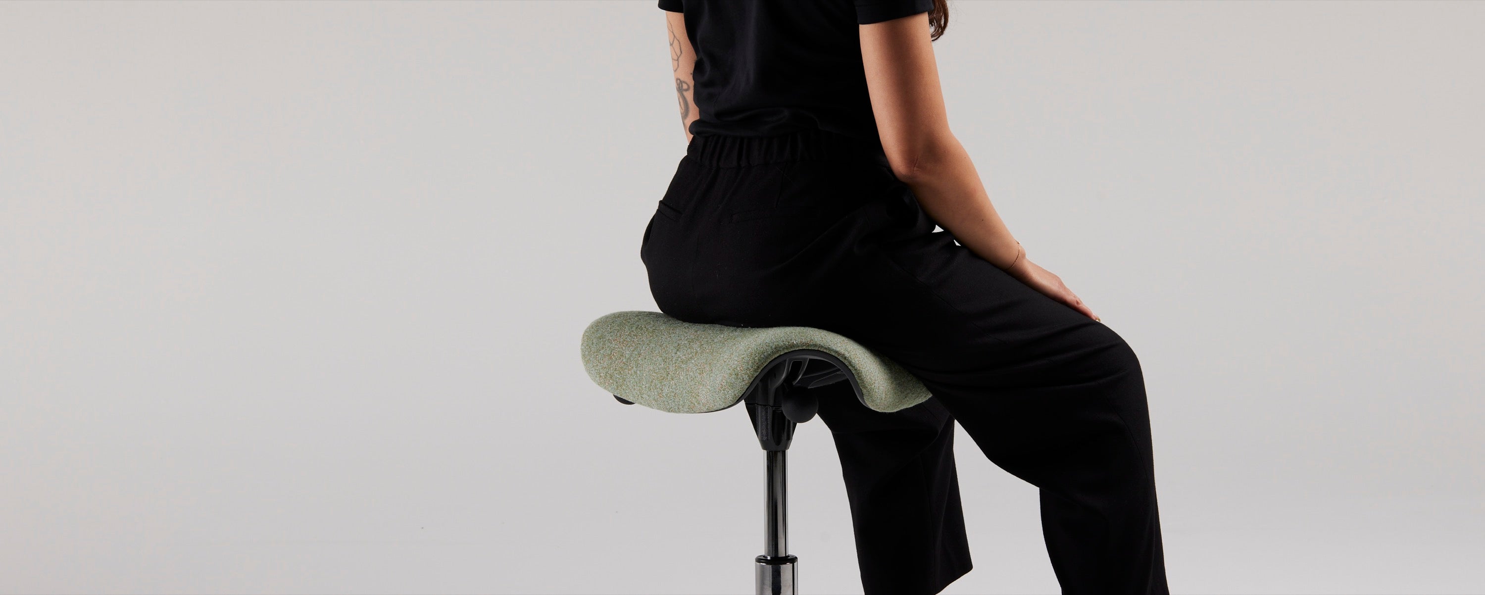 A rear view of a person sitting with an open-leg posture on a green Humanscale Freedom Saddle stool, showing the black frame and the healthy spinal alignment encouraged by the triangular seat