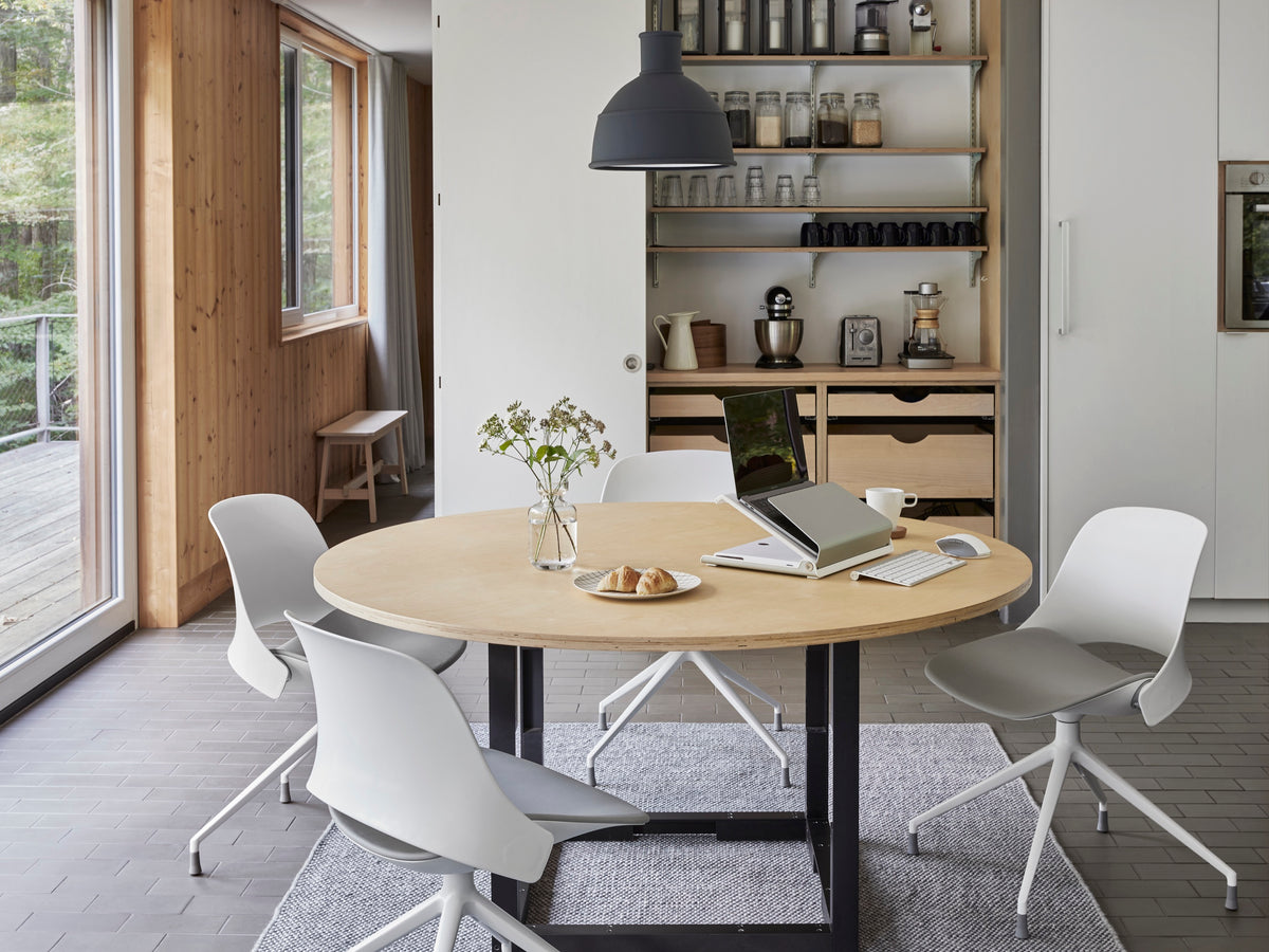 Four beige Humanscale Trea swivel chairs with casters around a wooden dining table featuring an L6 laptop holder notebook manager for an ergonomic home office setup