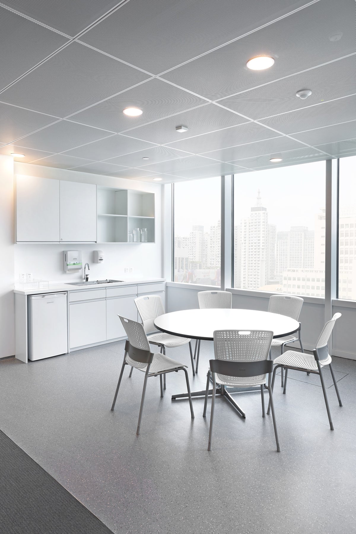 White and gray Humanscale Cinto armless chairs with perforated backs around a round white table in a modern office break room with a city view