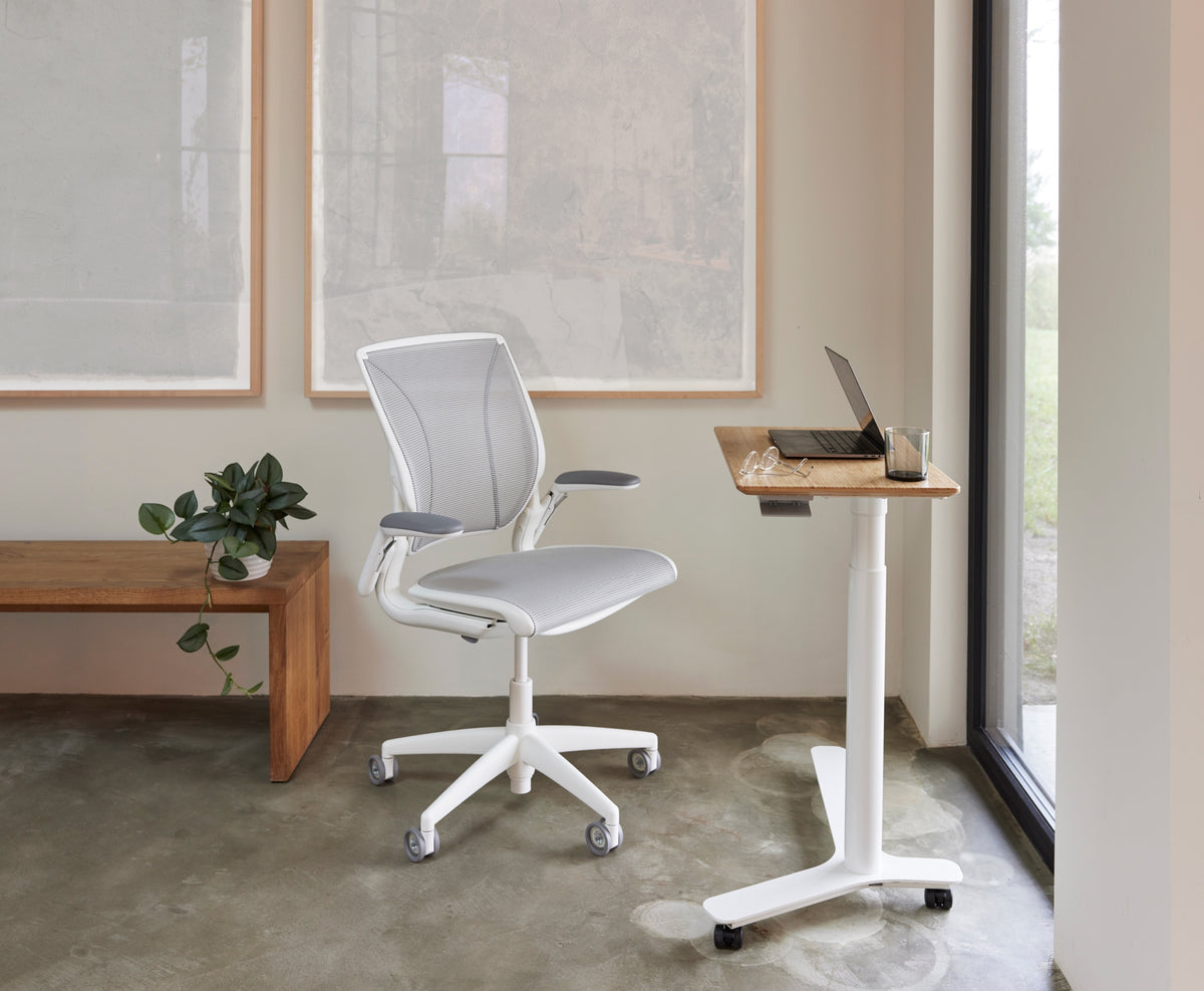 Lifestyle view of a Humanscale World Mesh chair in pinstripe white and a Float Mini standing desk with a bamboo surface and casters in a bright office