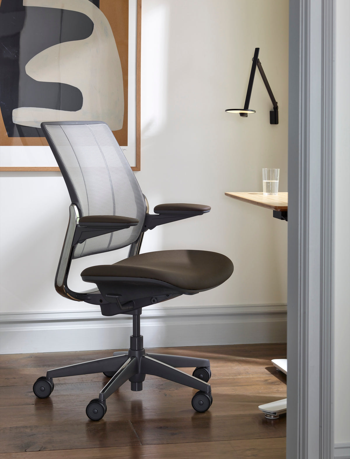 Side view of a Humanscale Diffrient Smart chair with black tri-paneled mesh, silver frame, and dark brown seat in a modern home office with a wooden desk and natural light