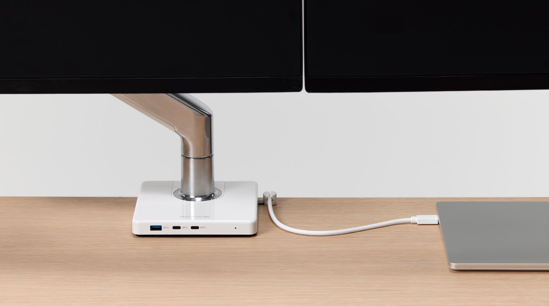 A close-up of a Humanscale M/Connect 3 centralized hub integrated into a monitor arm base, showing a silver laptop connected via a single cable for data and power on a light wood desk