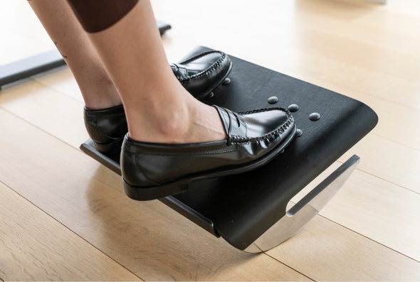 Feet resting in black leather loafers on Humanscale FR100 black adjustable footrest metal base